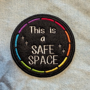 May include: A black and white embroidered patch with a rainbow border. The patch says "This is a safe space" with stars on either side of the word "safe".