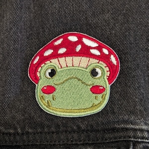 May include: Embroidered patch of a frog with a mushroom cap. The frog has a green face, red cheeks, and black eyes. The mushroom cap is red with white spots. The patch is attached to a black denim jacket.