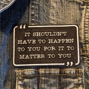 May include: A black rectangular patch with white text, reading "IT SHOULDN'T HAVE TO HAPPEN TO YOU FOR IT TO MATTER TO YOU." The patch is attached to a blue denim jacket, above a pocket.