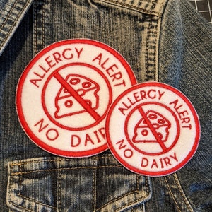 May include: Two white and red round allergy alert patches with a red line through a white cheese wedge. The text on the patches reads "Allergy Alert No Dairy". The patches are attached to a blue denim jacket.