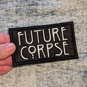 May include: A black rectangular patch with the words "FUTURE CORPSE" embroidered in white. The patch has a black border and is held against a light blue denim background.