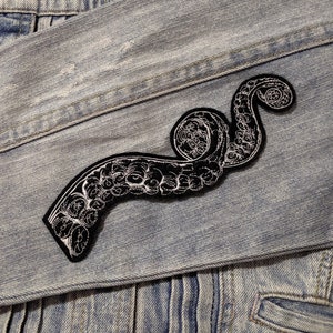 May include: A black embroidered patch of an octopus tentacle on a light blue denim jacket.