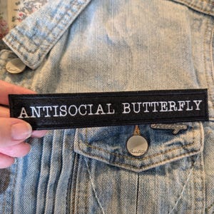 May include: A rectangular black fabric patch with white embroidered text that reads ANTISOCIAL BUTTERFLY. The patch is positioned horizontally against a light blue denim jacket featuring silver metal buttons and visible stitching details.