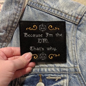 May include: Black felt patch with gold embroidered text that reads "Because I'm the DM. That's why." with a gold embroidered 20-sided die above and below the text.