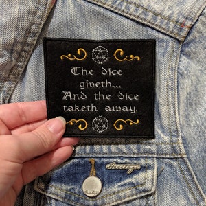 May include: Black fabric patch with gold embroidery. The patch features a stylized dice symbol and the text "The dice giveth... And the dice taketh away."