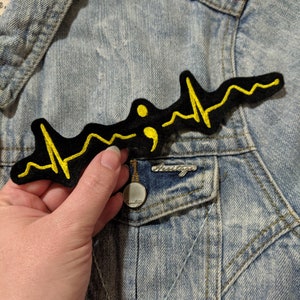May include: A black and yellow embroidered patch with a stylized heartbeat and a semicolon in the center. The patch is sewn onto a light blue denim jacket.
