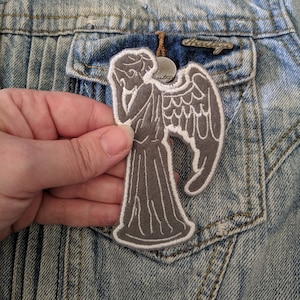 May include: A gray embroidered patch of a weeping angel from the Doctor Who television series. The patch is on a denim jacket.