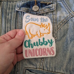 May include: A white felt patch with embroidered text that reads "Save the chubby unicorns". The patch features a yellow rhinoceros with a blue horn and a green outline.
