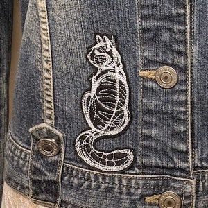 May include: A blue denim jacket with a white embroidered cat skeleton patch on the front.