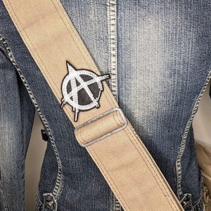May include: A tan guitar strap with a black and white anarchy symbol patch. The strap is worn over a denim jacket.