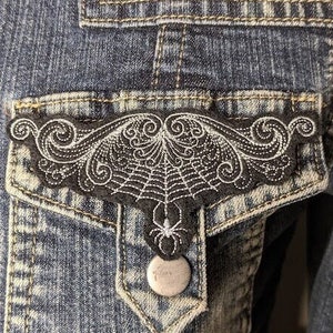 May include: A denim jacket with a black embroidered spiderweb and spider design on the pocket flap.