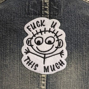 May include: A blue denim jacket with a white embroidered patch. The patch features a cartoon figure and the text "FUCK U THIS MUCH" in black, centered on the back.