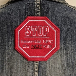 Peut inclure: Patch brodé rouge et blanc en forme de panneau d'arrêt. Le patch indique "STOP Essential NPC Do NOT Kill!" Le patch est cousu sur une veste en jean bleue.