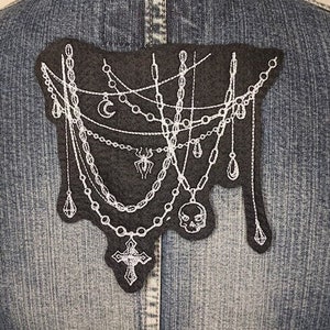 May include: A black felt patch with white embroidery of a spider, skull, cross, and crescent moon hanging from chains. The patch is sewn onto a denim jacket.