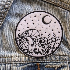 Peut inclure: Un écusson brodé blanc avec un contour noir d'un champignon, d'un croissant de lune et d'étoiles sur une veste en jean.