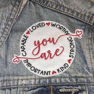 May include: White embroidered patch with red hearts and the text "You are" in red cursive. The words "Capable, Loved, Worthy, Strong, Important, Kind" surround the text in a circle.