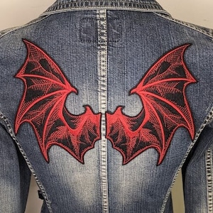 May include: A denim jacket with a large, embroidered bat wing design on the back. The wings are red with black detailing, creating a striking contrast against the blue denim. The jacket has a classic button-up design.
