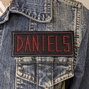 May include: A blue denim jacket with a black patch that says "DANIELS" in red lettering.