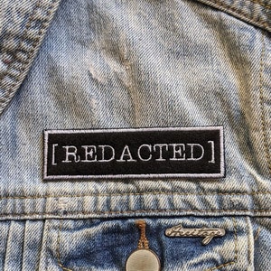 May include: A close-up of a denim jacket with a black patch that says "[REDACTED]" in white text. The jacket has a pocket with a metal button that says "Freitag" on it.