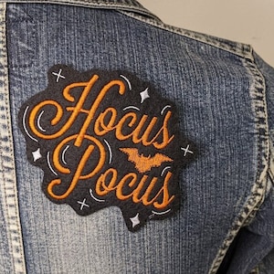 May include: A black and orange embroidered patch with the text "Hocus Pocus" in a cursive font. A small orange bat is centered above the text. The patch is sewn onto a blue denim jacket.