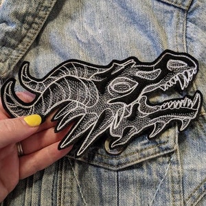 May include: Black and white embroidered dragon head patch. The patch is detailed with intricate stitching and is perfect for adding a unique touch to clothing or accessories.