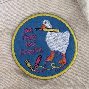 May include: An embroidered patch featuring a white goose holding a yellow crayon in its beak. The text "Be Gay Do Crafts" is embroidered in yellow on the patch, which is blue with a yellow border.