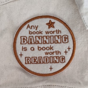 May include: A white embroidered patch with brown stitching. The patch is round and features the text "Any book worth banning is a book worth reading" with a star and plus signs.