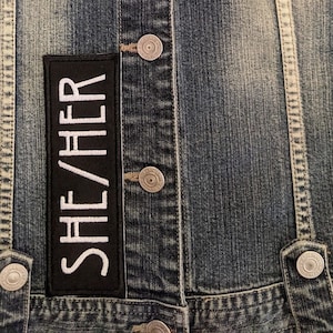 May include: A black denim jacket with a white patch that says "SHE/HER" on it.