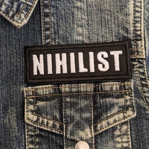 May include: Close-up of a denim jacket with a black patch that reads "NIHILIST" in white block letters. The jacket has a button-down front, two chest pockets, and silver buttons. The denim is a faded blue color.