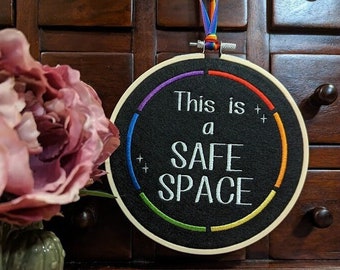 Könnte beinhalten: Ein runder Stickrahmen mit schwarzem Filzgrund und regenbogenfarbener Stickerei. Der Text "This is a SAFE SPACE" ist in Weiß auf den Filz gestickt.