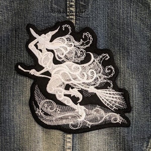 May include: White embroidered patch of a witch flying on a broomstick against a black background. The witch is depicted in a flowing gown with intricate details.