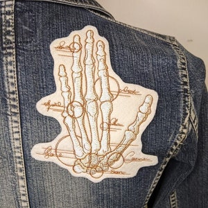 May include: A white denim jacket with a beige embroidered patch of a hand skeleton with anatomical details.