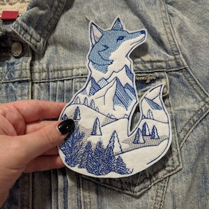 May include: Embroidered patch of a blue fox with a mountain range inside its silhouette. The fox is outlined in white and the mountains are filled with blue and white embroidery. The background is a light blue denim fabric.