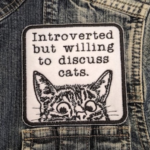 May include: A white patch with black embroidery that reads "Introverted but willing to discuss cats." A black and white illustration of a cat's face peeks out from the bottom of the patch.
