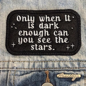 May include: A black embroidered patch with the quote, "Only when it is dark enough can you see the stars." The patch is rectangular with rounded corners and is sewn onto a denim jacket. The text is in white.