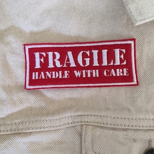 May include: Red rectangular fabric patch with white stitching. The patch says "FRAGILE HANDLE WITH CARE" in white letters.