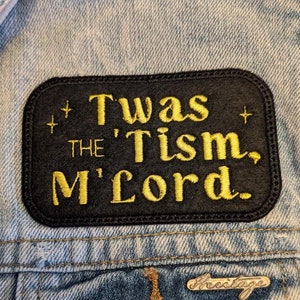 Peut inclure: Un écusson en tissu noir avec du texte brodé en or qui dit "Twas the 'Tism, M'Lord." L'écusson est cousu sur une veste en jean bleue.