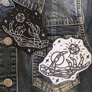 May include: Two embroidered iron-on patches featuring a skeletal hand holding a 20-sided die. The patch on the left is black with white stitching and the patch on the right is white with black stitching. Both patches have a star and crescent moon design.