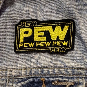 May include: A black and yellow embroidered patch with the word "PEW" in large yellow letters and the words "PEW PEW PEW" in smaller yellow letters below. The patch is sewn onto a blue denim jacket.