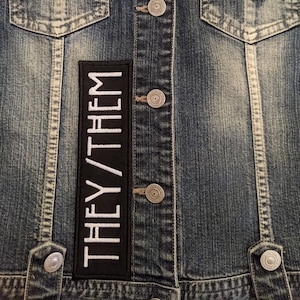 May include: A black denim jacket with a white embroidered patch that says "THEY/THEM" on the front.