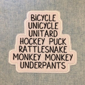 May include: A white embroidered patch with a light pink border. The patch features the words "BICYCLE, UNICYCLE, UNITARD, HOCKEY PUCK, RATTLESNAKE, MONKEY MONKEY, UNDERPANTS" in black, stacked text. The patch is on a light blue denim background.