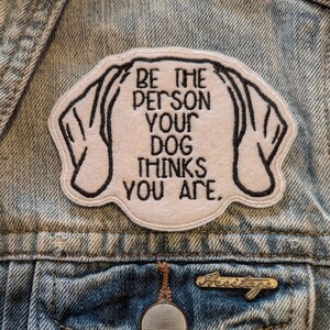 May include: A white patch with black stitching on a denim jacket. The patch says "Be the person your dog thinks you are."