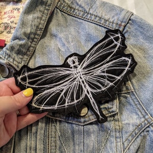 May include: A white embroidered dragonfly patch on a black felt background. The patch is attached to a blue denim jacket.