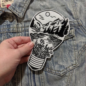 May include: Black and white embroidered iron-on patch of a lightbulb with a mountain scene inside. The scene includes a lake, mountains, trees, and a moon.