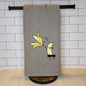 May include: A gray waffle-weave kitchen towel with a humorous cartoon banana design. The design features a banana with a black censor bar and a yellow banana peel. The towel is hanging on a black towel bar.