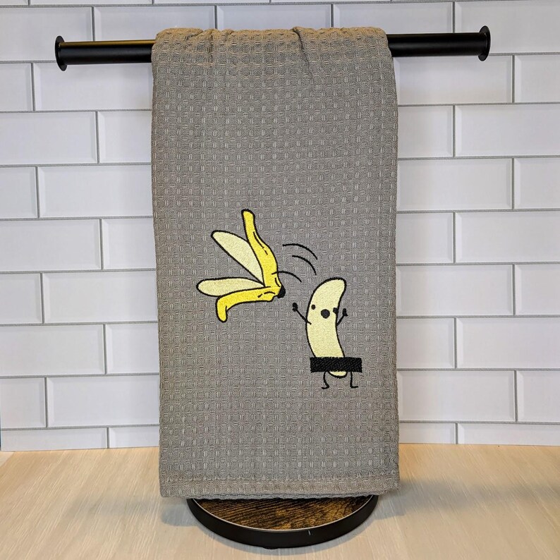 May include: A gray waffle-weave kitchen towel with a humorous cartoon banana design. The design features a banana with a black censor bar and a yellow banana peel. The towel is hanging on a black towel bar.