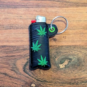 May include: Black lighter case featuring three green cannabis leaf designs. It has a silver keyring and a green accent. The lighter has a red button.