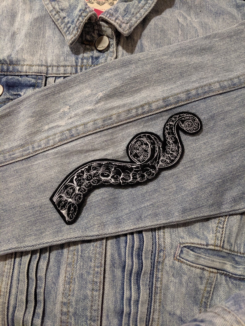 May include: A black and white embroidered octopus tentacle patch sewn onto a light blue denim jacket.
