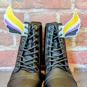 May include: A pair of black lace-up boots with non-binary pride flag wing-shaped patches attached to the top. The wings feature yellow, white, purple, and black colors. The boots are set against a brick wall background.