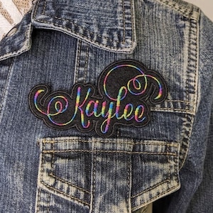 May include: A black and rainbow embroidered patch with the name "Kaylee" in a cursive font.